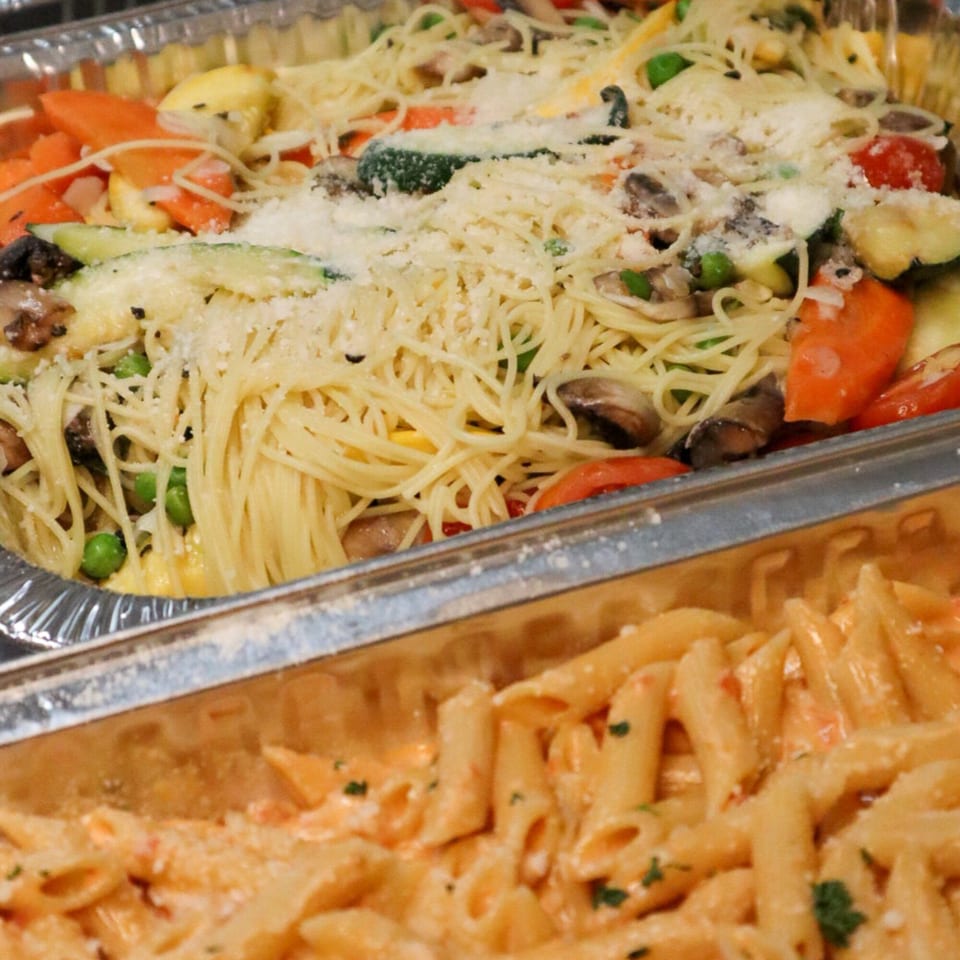 Two foil trays of pasta, one with spaghetti and vegetables, the other with penne in a creamy sauce.