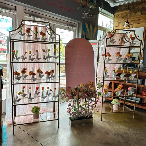 Two tall gold shelves filled with small vases of flowers, flanking a pink sign at Bacco Centro.