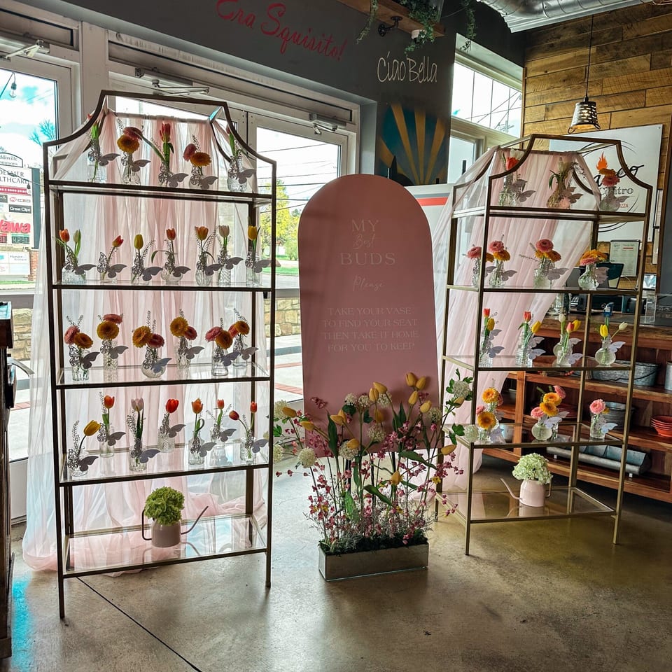 Two tall gold shelves filled with small vases of flowers, flanking a pink sign at Bacco Centro.