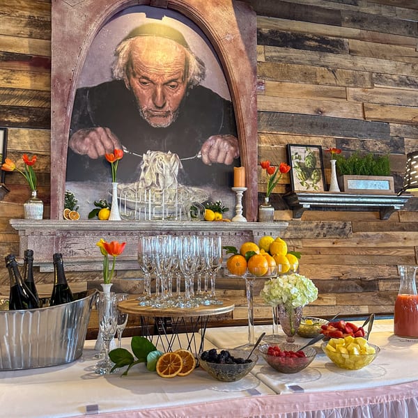 A Bacco Centro buffet table with fruit, champagne, and a large painting of a man eating pasta.