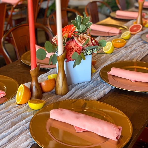 A Bacco Centro dining table set with gold plates, pink napkins, candles, flowers, and citrus halves.