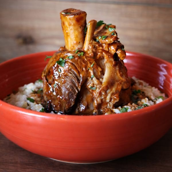 Braised lamb shank with rich sauce and fresh herbs, served over risotto in a red bowl.